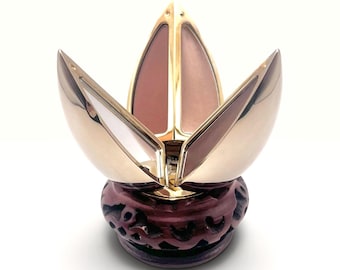 May include: A gold-toned, multi-petaled cosmetic compact that opens like a flower, revealing two distinct shades of pressed powder. The compact is supported by a dark, textured, circular base with intricate cut-out patterns along its side.