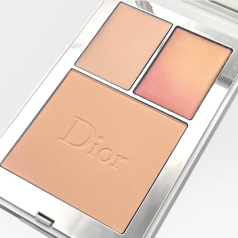 May include: A silver compact featuring a Dior face palette. The palette contains three shades: a large, light peach powder with the Dior logo, and two smaller shades of peach and a peach-pink gradient.