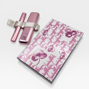 May include: A pink makeup set featuring a mascara and a lipstick, held together with a silver band. A matching compact with a floral and logo pattern is also present. The set is on a white background.