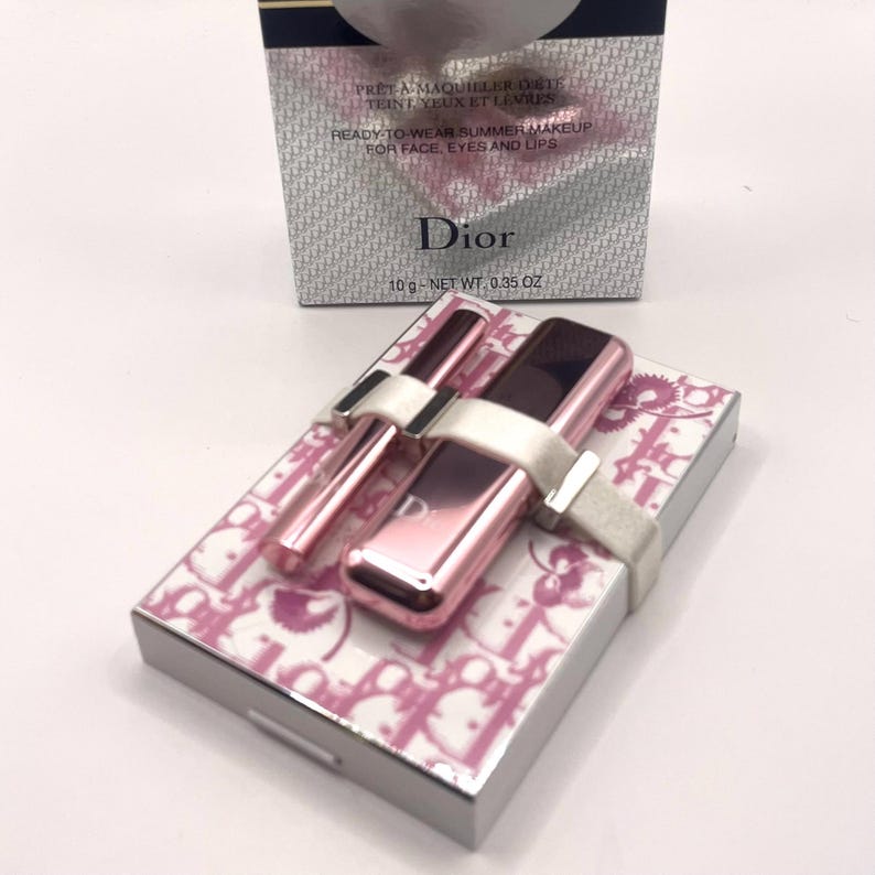 May include: A Dior makeup set featuring a pink lipstick, a brush, and a compact. The compact and lipstick are pink with the Dior logo. The set is tied with a white ribbon and presented on a box with a pink and white pattern. The box reads "READY-TO-WEAR SUMMER MAKEUP FOR FACE, EYES AND LIPS".