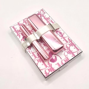 May include: A pink makeup set featuring a lipstick and mascara, secured with a white ribbon. The set rests on a patterned box with a pink and white design. The lipstick and mascara are a shiny pink color.