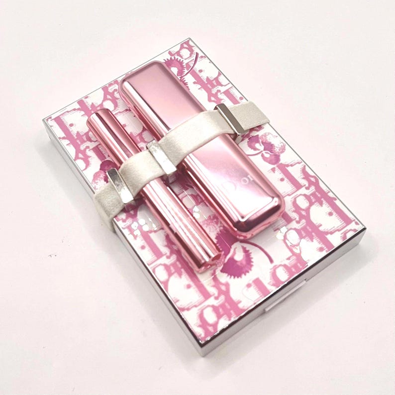 May include: A pink makeup set featuring a lipstick and mascara, secured with a white ribbon. The set rests on a patterned box with a pink and white design. The lipstick and mascara are a shiny pink color.