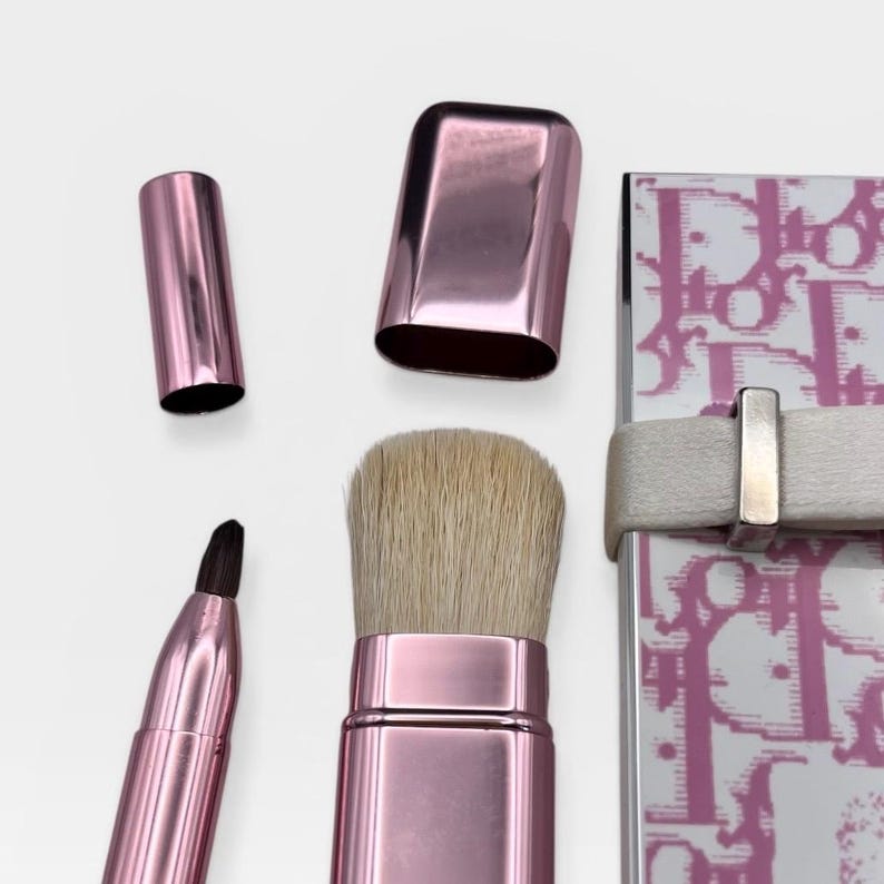 May include: A set of pink makeup brushes and a compact. The brushes include a large powder brush, a small angled brush, and a liner brush. The compact is white with a pink pattern and a white strap.