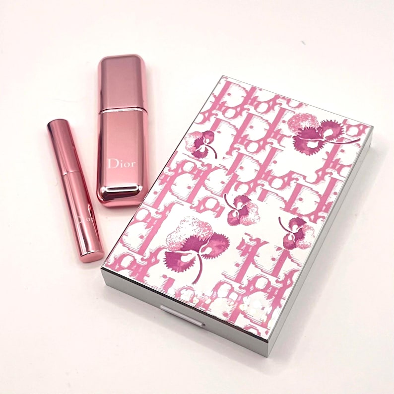 May include: A collection of pink Dior beauty products. Includes a mascara tube, a perfume atomizer, and a rectangular compact with a white and pink floral pattern and the Dior logo.