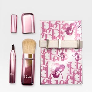 May include: A collection of pink Dior makeup brushes and accessories. Includes a brush with a cap, a small brush, and a compact with a floral pattern and a white band. The Dior logo is visible on the brushes.