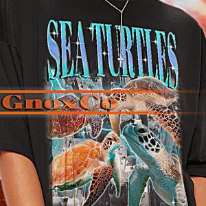 Könnte beinhalten: Schwarzes Unisex-T-Shirt mit einem farbenfrohen Seeschildkröten-Motiv und dem Schriftzug "SEA TURTLES" in Türkis. Das Oberteil trägt den Markennamen "TSHIRT Unisex". Lässiger, übergroßer Stil.