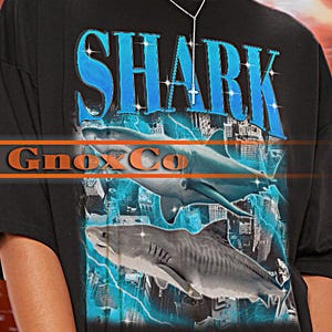 Könnte beinhalten: Schwarzes Unisex-T-Shirt mit einem Hai-Motiv. Das Wort "SHARK" ist blau, mit einem Hai-Bild und dem Text "Gnox Co" darunter. Das T-Shirt hat kurze Ärmel und eine lockere Passform. Der Hintergrund ist ein verschwommenes rotes und oranges Blitzdesign.
