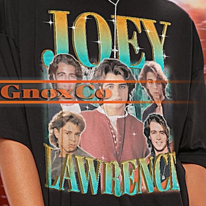 May include: Black unisex t-shirt featuring the text "JOEY LAWRENCE" above a collage of images. The shirt has a crew neck and short sleeves. The brand name "TSHIRT Unisex" is at the top left.