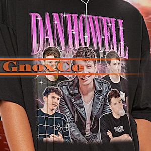 May include: Black unisex t-shirt featuring the text "DAN HOWELL" in a purple font, with a photo of five people. The shirt also has the text "TSHIRT Unisex" at the top. The model is wearing the shirt.