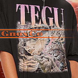 May include: Black unisex T-shirt with the word "TEGU" in pink letters. The shirt features a graphic of lizards and the text "Gnoxica". The T-shirt is part of the TSHIRT Unisex collection.