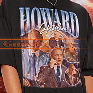 May include: Black unisex t-shirt with the text "HOWARD" in a blue and white design, and a collage of images. The shirt has short sleeves and a crew neck. The brand name "TSHIRT Unisex" is in the upper left corner.
