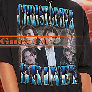Könnte beinhalten: Schwarzes Oversize-T-Shirt mit einem Band-Grafikdruck von "Christopher Briney" mit dem Text "Gnox Co" in der Mitte. Das Model trägt das Shirt mit weißen Shorts und einer schwarzen Sonnenbrille.