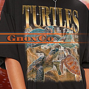Könnte beinhalten: Schwarzes Unisex-T-Shirt mit goldenem "TURTLES"-Aufdruck und Schildkröten-Illustrationen. Das Shirt hat auch den Text "Gnox Co" und "TSHIRT Unisex". Das Model trägt Größe 2XL.