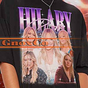 Könnte beinhalten: Schwarzes Unisex-T-Shirt mit einem Grafikdruck von Hilary Duff. Das Shirt hat einen Vintage-Look mit einem lila und pinkfarbenen Farbschema. Der Text "Hilary Duff" ist in einer großen, fetten Schrift gedruckt. Das Model trägt ein Shirt in Größe 2XL.