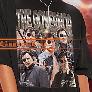 May include: Black t-shirt with a graphic print of a group of people with the text "THE GOVERNOR" above them. The t-shirt is oversized and has a distressed look.