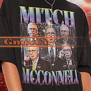 Puede incluir: Camiseta unisex negra con un estampado gráfico de un grupo de caras de hombres. El texto "Mitch McConnell" está impreso en la camiseta. La modelo lleva una camiseta talla 2XL.