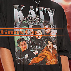 May include: Black oversized t-shirt featuring a band graphic print of "Katy O'Brian" with the text "Gnox Co.". The model is wearing a white vest top underneath the t-shirt.