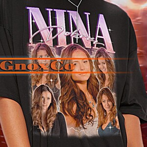 Puede incluir: Camiseta negra de gran tamaño con un estampado gráfico de la cara de una mujer y el texto "NINA" en estilo vintage. La camiseta la lleva puesta una persona con pantalones cortos blancos.