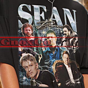 May include: Black unisex t-shirt featuring the text "SEAN" in large, bold letters above a collage of images. The shirt also has the text "Gnoxu" in red. The model is wearing sunglasses. The shirt is true to size.