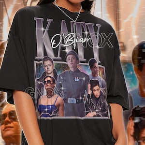 May include: Black unisex t-shirt with a graphic design featuring the text "KATY O'Brian" and images of people. The shirt also has the text "GNOX" and "TSHIRT Unisex". The model wears a 2XL shirt.