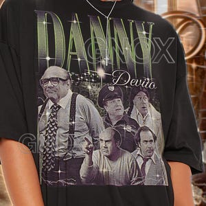 May include: Black unisex t-shirt featuring a graphic of Danny DeVito with the text "DANNY DeVito". The design includes images of DeVito in various roles. The shirt has a vintage style and is labeled "TSHIRT Unisex".