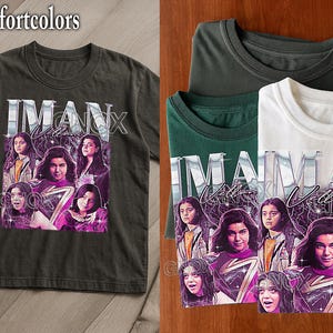 May include: Dark grey Comfort Colors t-shirt featuring a graphic of a woman's face with the text "IMAN Vellani". The design includes a purple and white lightning bolt and multiple images of the woman. Several folded t-shirts are also visible.