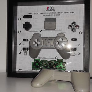 Op de afbeelding: Een ingelijst display van een gedemonteerde Sony PlayStation-controller met de tekst "SONY PLAYSTATION CONTROLLER SCPH-1001 DECEMER 5, 1994" en "P-30 Anniversary". Een grijze PlayStation-controller bevindt zich op de voorgrond.