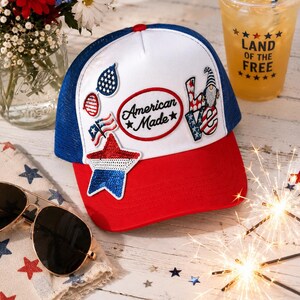 May include: A white and blue trucker hat with a red brim, featuring a central oval patch that reads American Made. The hat is decorated with various patriotic patches, including a sequined star, a gnome, a flag, and glasses in red, white, and blue.