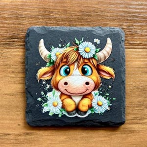May include: Square black slate coaster featuring a cartoon Highland cow with a floral crown and daisies. The cow has brown fur, large blue eyes, and a white muzzle. The coaster is approximately 10 cm square.