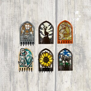 May include: A collection of six decorative key holders, each with an arched design. The key holders feature a pig, deer, fish, anchor, sunflower, and cow. They are black and have multiple hooks for keys.