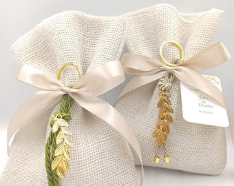 Puede incluir: Dos bolsas de arpillera en color blanco roto atadas con cintas de satén color champán. Cada bolsa presenta un dije de hoja de metal dorado y una borla de cordón trenzado. Una etiqueta blanca muestra el texto "John & Dimitra 30.08.2025".