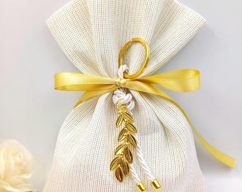 Puede incluir: Bolsa de regalo de tela blanca con brillo dorado, atada con una cinta de satén amarilla y un dije de rama de olivo dorado.