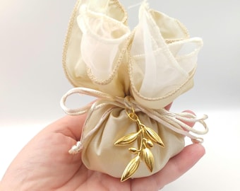 May include: A small, beige satin pouch with a drawstring closure, topped with a ruffled, sheer fabric. A gold-colored olive branch charm hangs from the pouch. The pouch is held in a hand.