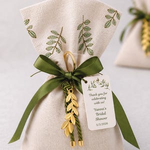 May include: A beige linen gift bag tied with a green satin ribbon and a gold leaf charm. The bag has embroidered green leaves and a tag that reads "Thank you for celebrating with us! Yanna's Bridal Shower 5/3/2026."