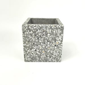 Könnte beinhalten: Quadratischer grauer Pflanztopf mit einem gesprenkelten Terrazzo-Design. Der Pflanztopf hat eine glatte, polierte Oberfläche und eine schlichte, moderne Ästhetik. Das gesprenkelte Muster umfasst weiße, schwarze und beige Flecken.