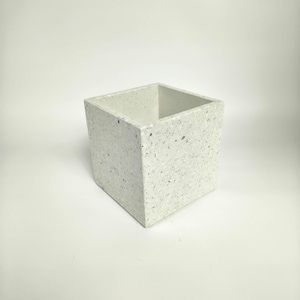 Può includere: Un vaso di forma quadrata con una base di colore bianco e un motivo in stile terrazzo caratterizzato da piccole macchie grigie e nere su tutta la superficie esterna. Il contenitore presenta un interno cavo e una parte superiore piatta.