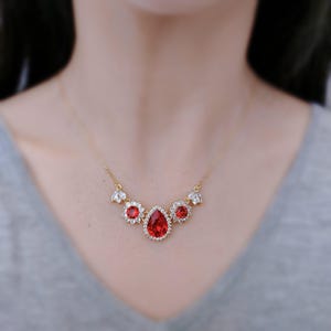 Op de afbeelding: Een gouden ketting met een traanvormige rode strass-hanger omringd door kleinere heldere strass-stenen. De ketting heeft aan weerszijden van de hanger twee kleinere ronde rode strass-stenen.