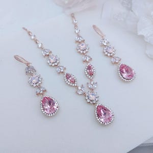 Può includere: Un set di gioielli in oro rosa con collana e orecchini abbinati. Il set è adornato con pietre preziose rosa a forma di goccia e floreali, rifinite con cristalli trasparenti. La collana e gli orecchini sono esposti su una superficie bianca.