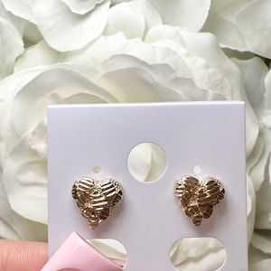 May include: A pair of gold heart-shaped earrings with a textured surface. The earrings are displayed on a white card with two oval cutouts.