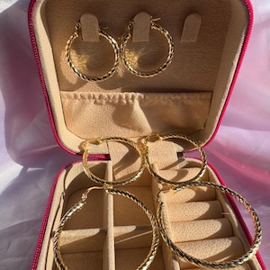 Puede incluir: Cuatro pares de pendientes de aro dorados con un diseño de cuerda trenzada texturizada. Los pendientes se exhiben dentro de un estuche de joyería rosa con interior beige. El estuche está abierto, mostrando los pendientes.