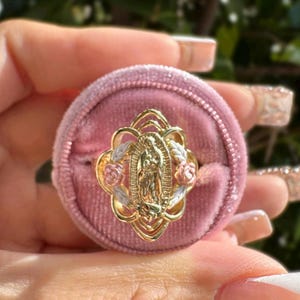 May include: Gold-toned ring featuring the Virgin Mary, surrounded by floral and leaf accents. The ring is set in a pink velvet ring box. The ring's design includes rose and leaf details, creating a religious and decorative piece.