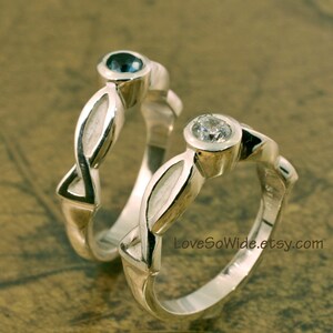 May include: Two silver rings with intertwined bands. One ring has a blue gemstone and the other has a clear gemstone. The rings are on a brown surface. LoveSoWide.etsy.com