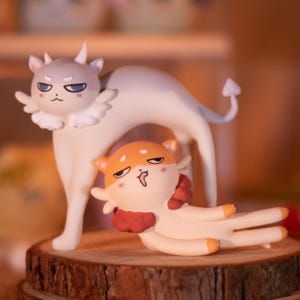 以下が含まれることがあります： 2匹の猫のような生き物を特徴とする気まぐれな置物セット。1つは白い体に灰色の頭と角があり、もう1つはオレンジとクリーム色です。どちらも翼と悪魔のような表情をしており、木の台座に置かれています。