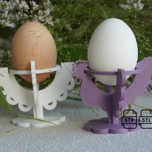 Op de afbeelding: Twee 3D-geprinte eierrekjes, een wit en een paars, in de vorm van een kip. Het witte rekje heeft een gegolfde rand. Beide rekjes bevatten een ei.