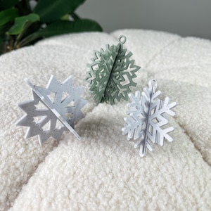May include: Three snowflake ornaments in shades of white and green. The ornaments are made of a lightweight material and have a three-dimensional design. The snowflakes are displayed on a white, textured surface. Perfect for winter holiday decor.