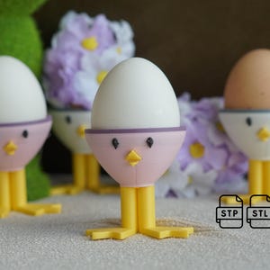 Puede incluir: Tres soportes para huevos impresos en 3D con forma de pollito con detalles en morado y rosa. Cada soporte tiene un huevo blanco en su interior. Los soportes están sobre una superficie blanca con flores moradas.
