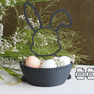 Könnte beinhalten: Schwarzer, metallener Osterhasen-Eierhalter mit einer schwarzen Schale und weißen und braunen Eiern darin. Der Halter steht auf einer weißen Oberfläche mit grünen Pflanzen im Hintergrund. Der Text "STP" und "STL" ist in der unteren rechten Ecke des Bildes sichtbar.