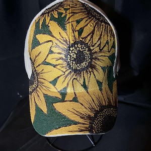 以下が含まれることがあります： 緑と黄色のひまわり柄の白いベースボールキャップ。ひまわりは、茶色の芯と黄色の花びらでリアルなスタイルで描かれています。