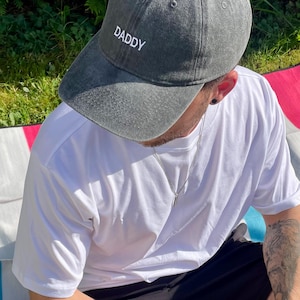Könnte beinhalten: Graue Baseballkappe mit dem Wort "DADDY" in Weiß gestickt. Die Kappe wird von einer Person getragen, die ein weißes T-Shirt und eine silberne Halskette trägt. Die Person sitzt draußen.