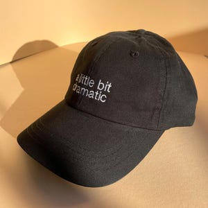 May include: Black baseball cap with the embroidered text "a little bit dramatic" in white. The cap has a curved brim and a classic six-panel design. The hat is made of a soft, durable fabric.